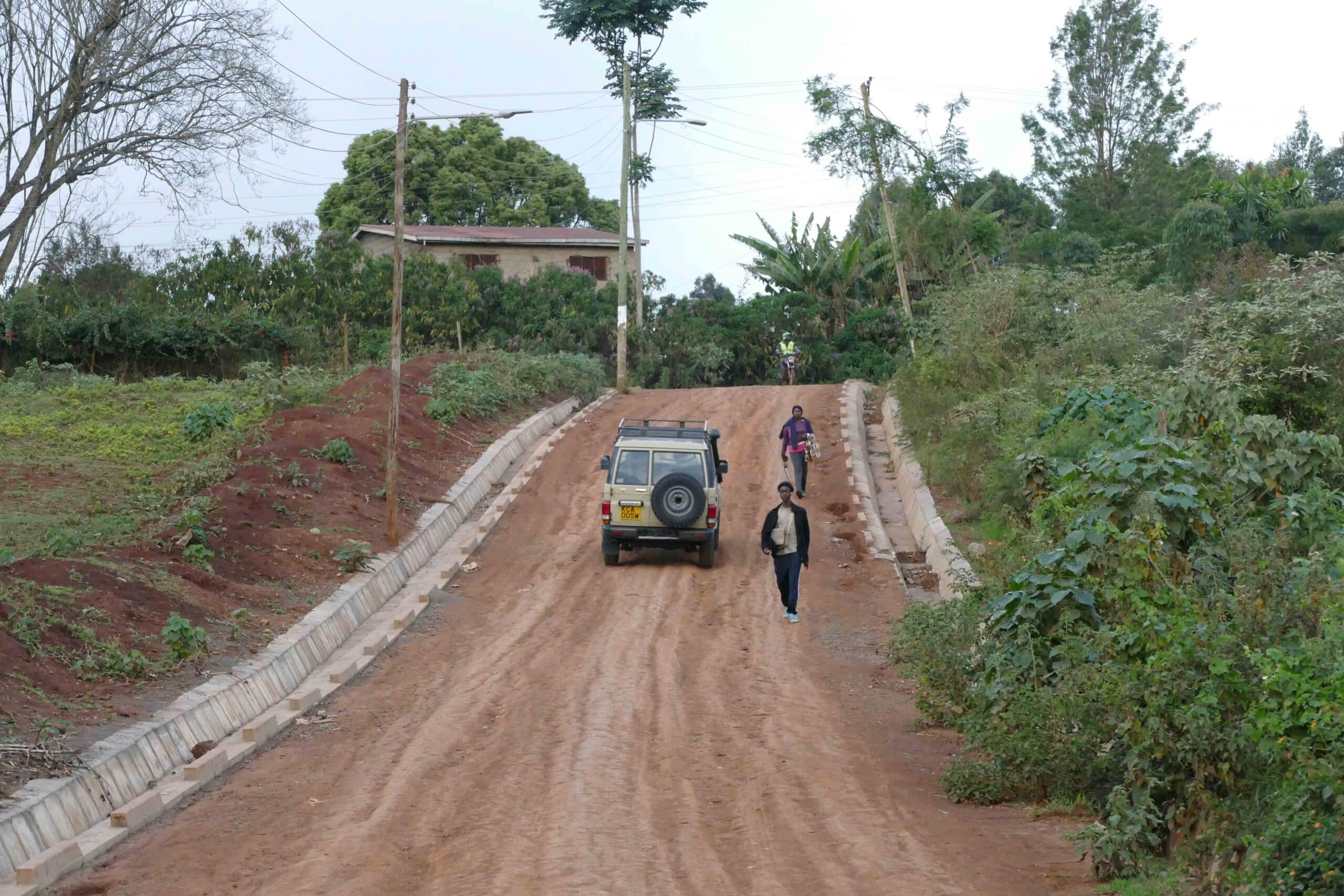 image shows a car on a dirt road with green vergetaion to the sides of the road